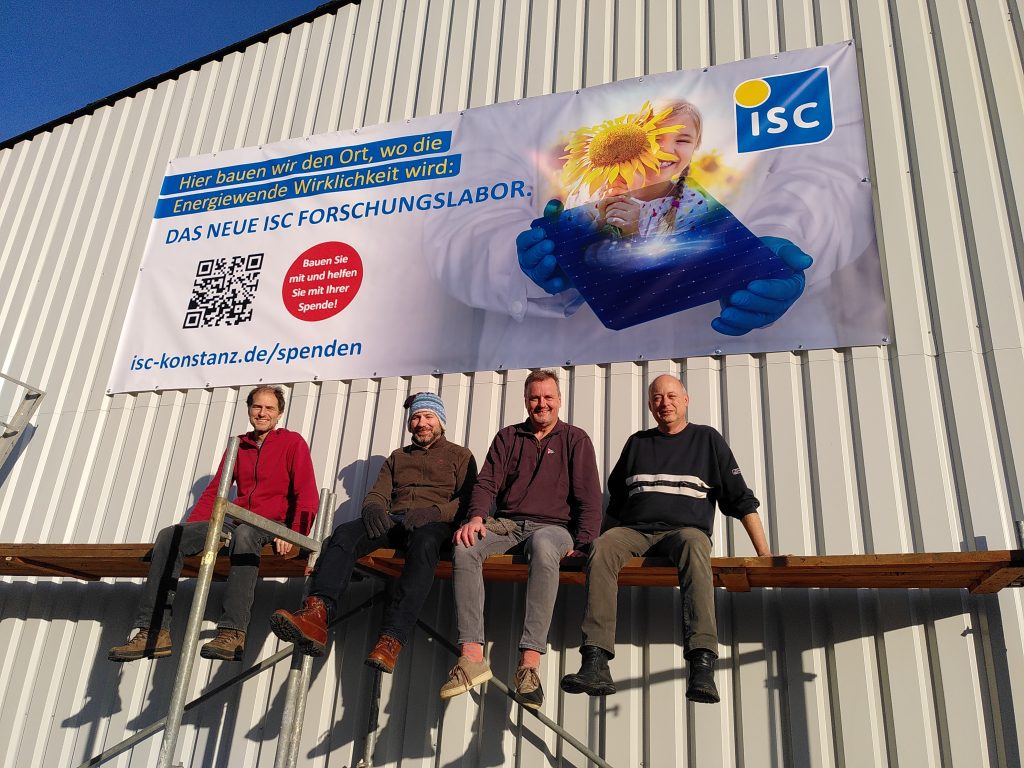 A banner with an appeal for donations (isc-konstanz.de/en/donations) hangs on a hall behind four men. The men are sitting on a scaffolding.